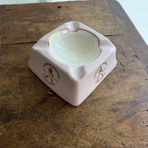 K&ouml;nnte beinhalten: Ein quadratischer Vintage-Aschenbecher in zartem Rosa. Der Aschenbecher hat eine cremefarbene Innenseite und einen goldenen Rand. Die Worte "Johnnie Walker Scotch Whisky" sind in Gold auf den Seiten aufgedruckt.
