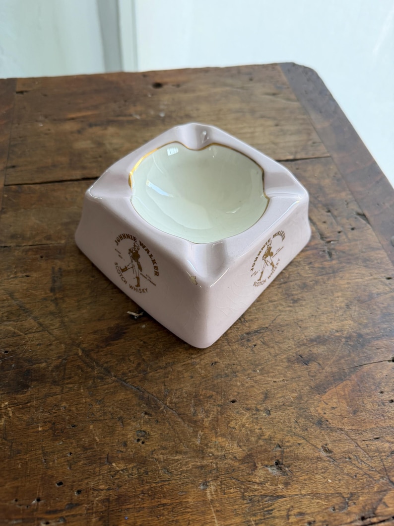 K&ouml;nnte beinhalten: Ein quadratischer Vintage-Aschenbecher in zartem Rosa. Der Aschenbecher hat eine cremefarbene Innenseite und einen goldenen Rand. Die Worte "Johnnie Walker Scotch Whisky" sind in Gold auf den Seiten aufgedruckt.