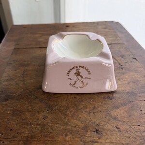 K&ouml;nnte beinhalten: Ein rosa Aschenbecher aus Keramik mit wei&szlig;em Innenraum und goldenem Rand. Der Aschenbecher tr&auml;gt das Logo "Johnnie Walker Scotch Whisky" in Gold und Schwarz. Der Aschenbecher ist rechteckig mit abgerundeten Kanten und steht auf einer Holzoberfl&auml;che.