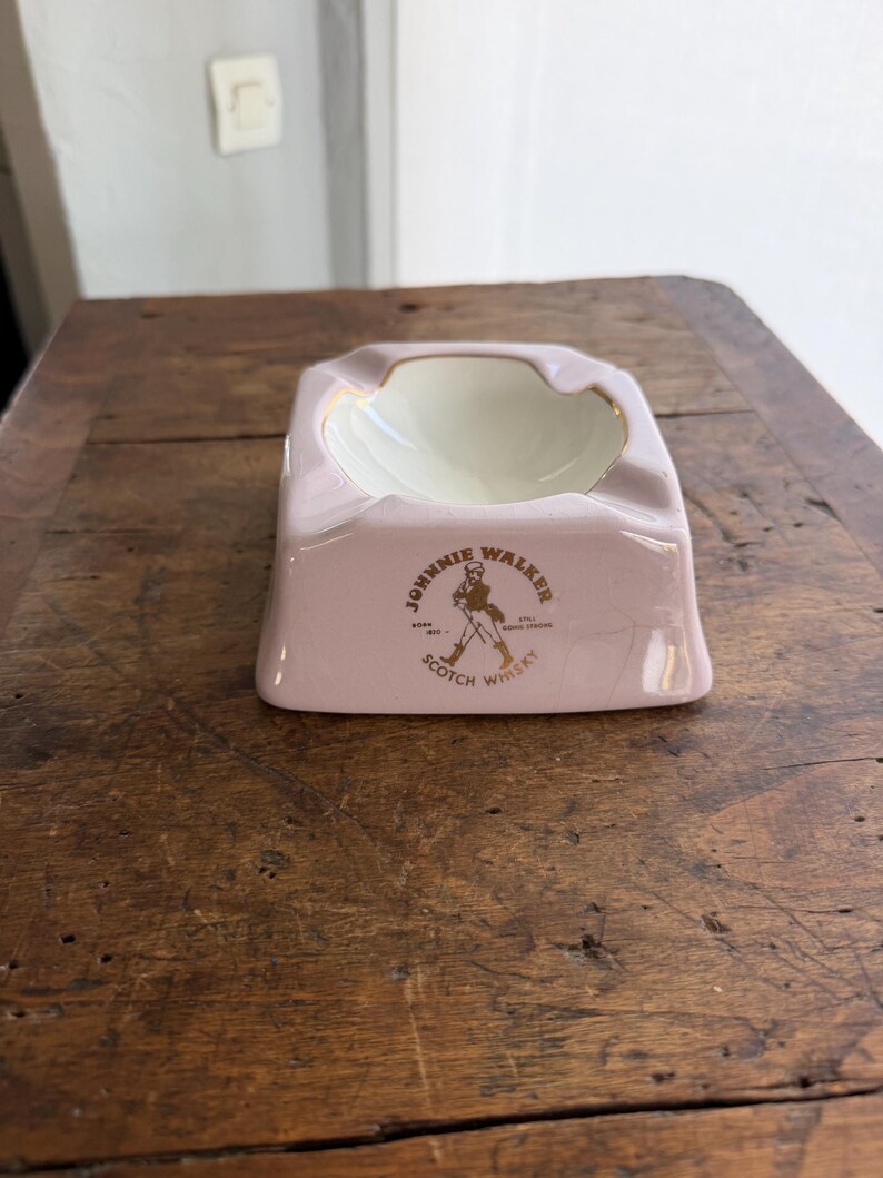 K&ouml;nnte beinhalten: Ein rosa Aschenbecher aus Keramik mit wei&szlig;em Innenraum und goldenem Rand. Der Aschenbecher tr&auml;gt das Logo "Johnnie Walker Scotch Whisky" in Gold und Schwarz. Der Aschenbecher ist rechteckig mit abgerundeten Kanten und steht auf einer Holzoberfl&auml;che.