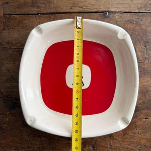 May include: Square ashtray with a white border and a red center with a white square and a black graphic. The ashtray measures approximately 7.5 inches across. Four small rests are on the sides.