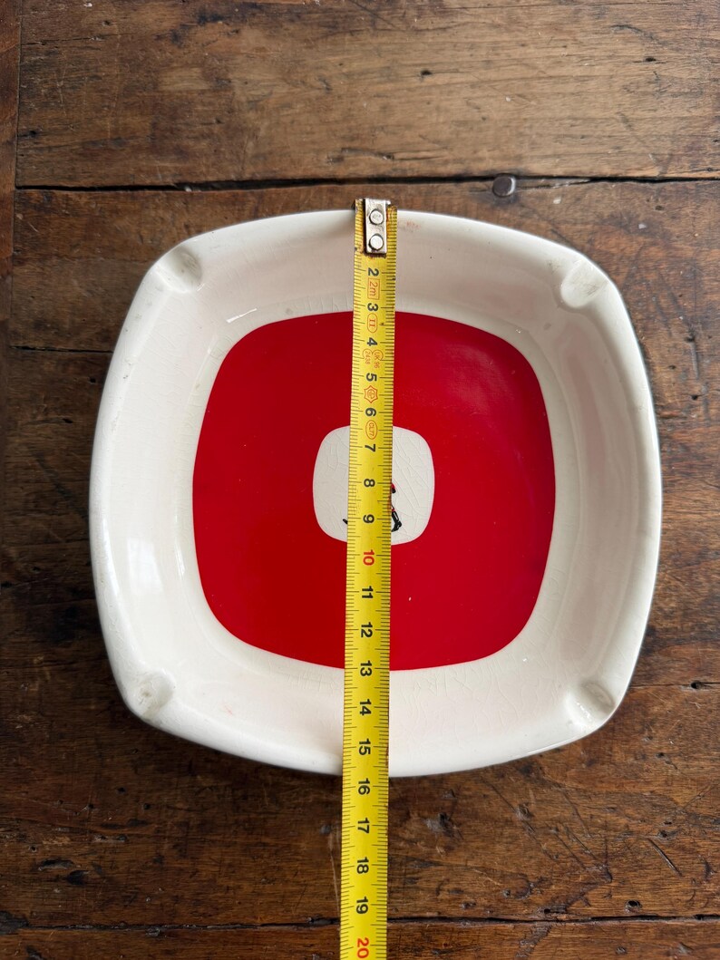 May include: Square ashtray with a white border and a red center with a white square and a black graphic. The ashtray measures approximately 7.5 inches across. Four small rests are on the sides.