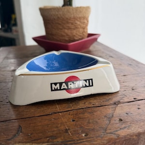 May include: A triangular ashtray with a white exterior, blue interior, and gold trim. The ashtray features the word "MARTINI" in black letters on a red circle. A plant in a woven pot sits in the background.