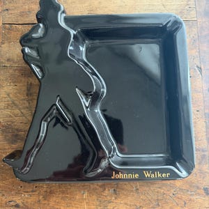 Pode incluir: Uma bandeja retangular preta brilhante com o logotipo Johnnie Walker em letras douradas. A bandeja apresenta uma silhueta em relevo da ic&ocirc;nica figura do Homem a Caminhar no lado esquerdo.