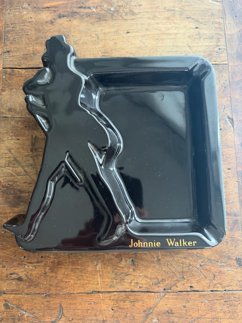 Pode incluir: Uma bandeja retangular preta brilhante com o logotipo Johnnie Walker em letras douradas. A bandeja apresenta uma silhueta em relevo da ic&ocirc;nica figura do Homem a Caminhar no lado esquerdo.