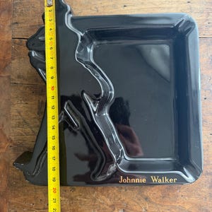 Pode incluir: Bandeja retangular preta com uma silhueta em relevo de um homem andando, com o texto "Johnnie Walker" em dourado. A bandeja mede aproximadamente 23 cm de comprimento.