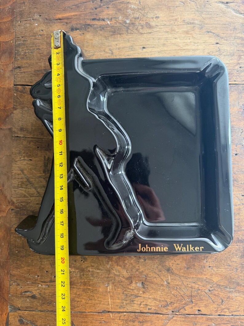 Pode incluir: Bandeja retangular preta com uma silhueta em relevo de um homem andando, com o texto "Johnnie Walker" em dourado. A bandeja mede aproximadamente 23 cm de comprimento.