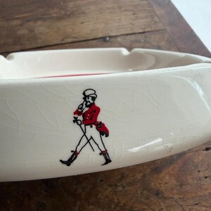 May include: Cream-colored ceramic ashtray with a red interior. The ashtray features a black and red illustration of a man in a red coat and black boots. The ashtray is on a wooden surface.