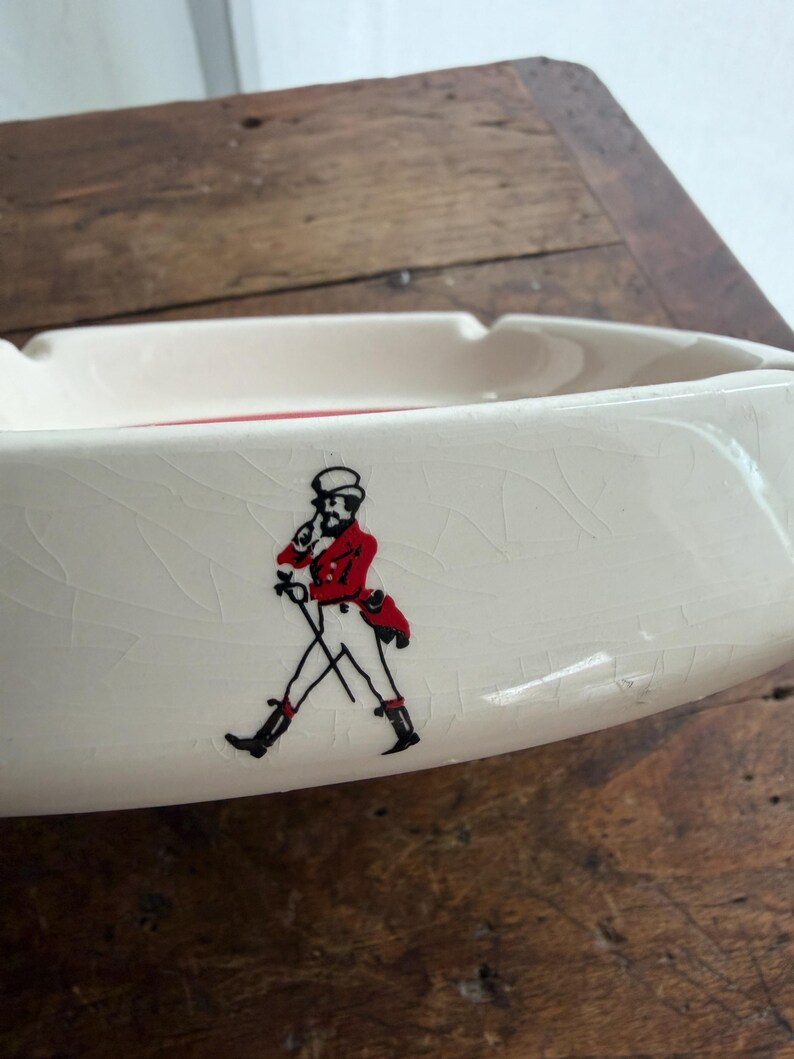 May include: Cream-colored ceramic ashtray with a red interior. The ashtray features a black and red illustration of a man in a red coat and black boots. The ashtray is on a wooden surface.
