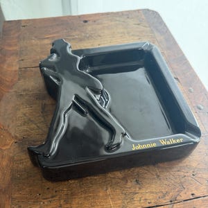 Pode incluir: Cinzeiro preto com formato retangular e silhueta em relevo de uma figura em movimento. As palavras "Johnnie Walker" est&atilde;o em letras douradas. O cinzeiro est&aacute; sobre uma superf&iacute;cie de madeira.