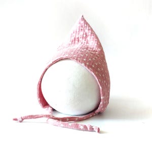 Op de afbeelding: Een lichtroze babykapje met een wit sterrenpatroon. Het kapje is gemaakt van zachte stof en heeft een puntige bovenkant en strikjes om het onder de kin vast te zetten. Het kapje wordt getoond op een witte, afgeronde vorm.