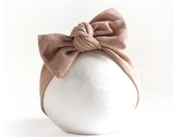 May include: A dusty rose headband featuring a large, knotted bow detail. The fabric has a subtle, textured pointelle pattern with small, repeating diamond-shaped perforations throughout the material, creating a soft and uniform appearance.