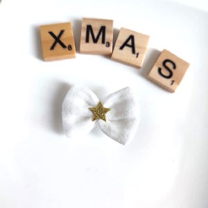 Könnte beinhalten: Ein weißer Stoff-Haar-Schleife mit einem goldenen Glitzerstern in der Mitte. Das Wort "XMAS" wird mit Holzbuchstaben geschrieben. Ein festliches Accessoire.
