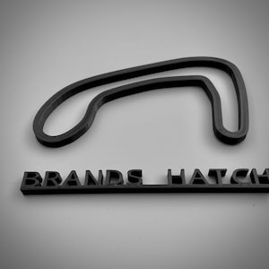 May include: A black 3D printed model of the Brands Hatch race track, a popular motorsports circuit in England. The model is displayed on a white surface with the name "Brands Hatch" printed below it.