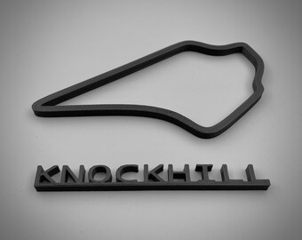 以下が含まれることがあります： スコットランドの有名なモータースポーツサーキット、ノックヒル・レースウェイの黒色の3Dプリントモデル。コースはシンプルなアウトラインで表示され、その下に「ノックヒル」という名前が印刷されています。