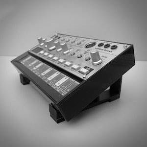 May include: A black and silver Volca Bass synthesizer with multiple knobs, buttons, and a small display. The device is angled upwards on a black stand. The text "volca bass" is visible on the top panel.