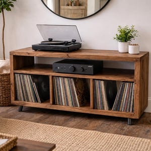 May include: A wooden media console with a black turntable, amplifier, and vinyl records. The console has three storage compartments filled with vinyl records. The unit is made of light brown wood and has black metal legs. Decorative plants sit on top.