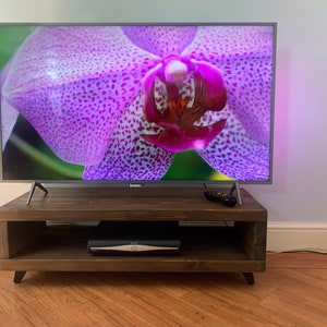 May include: A dark brown wooden TV stand with a shelf and black metal legs. A flat screen TV is on top of the stand, displaying a close-up image of a pink orchid.