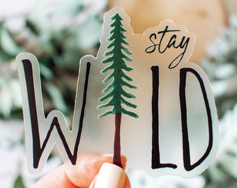 May include: A clear sticker with the text "stay wild" in black script. A green pine tree is in the center of the sticker. The sticker is slightly translucent.