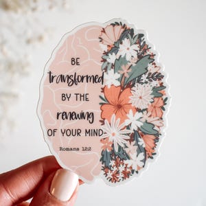 May include: A sticker with a brain design, half filled with flowers and foliage in shades of peach, white, and green. The other half of the brain is a solid peach color. The sticker has the words "Be transformed by the renewing of your mind" and "Romans 12:2".