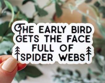 May include: White sticker with black text that reads "The early bird gets the face full of spider webs".  Two small pine tree graphics are on either side of the text.