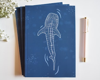 May include: A set of three blue notebooks with a white outline of a whale shark on the front cover. The notebooks have a speckled blue background.