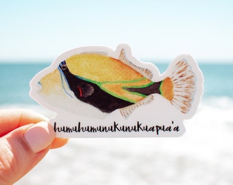 Könnte beinhalten: Ein weißer Aufkleber mit einer Aquarell-Illustration eines gelben, orangefarbenen, schwarzen und grünen Fisches. Der Fisch ist ein Drückerfisch und der Aufkleber trägt den Text "humuhumunukunukuapua'a" unter dem Fisch.