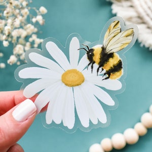 May include: A clear sticker with a white daisy and a yellow and black bumblebee. The bumblebee is perched on the daisy.