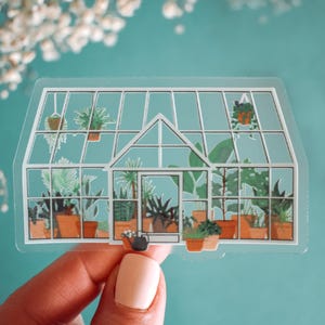 May include: A clear sticker of a greenhouse filled with various plants and a watering can. The sticker is on a blue background.