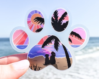 May include: A paw print shaped sticker with a sunset scene and two silhouettes of people and a dog on a beach. The sticker is on a blue and white background.