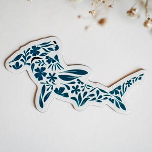 May include: A sticker featuring a hammerhead shark design. The shark is filled with dark teal floral patterns, including flowers and leaves, against a white background. The sticker has a white border and is likely for decorative purposes.