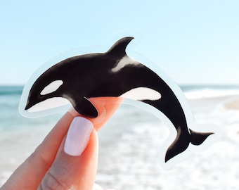 May include: A clear sticker of a black and white orca whale jumping out of the water. The sticker is on a person's finger, with the ocean in the background.