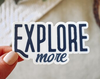 May include: A white sticker with the words "EXPLORE" in navy blue block letters above the word "more" in a navy blue script font. The sticker is held by a person.