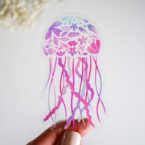 May include: A clear sticker featuring a jellyfish design with floral details. The jellyfish's bell is adorned with purple, pink, and blue flowers and leaves. Long, flowing tentacles in shades of pink and purple extend downward, creating a whimsical and artistic aesthetic.