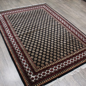 May include: A black and cream patterned rug with a red and white border. The rug has a fringe around the edges.
