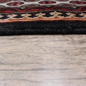 May include: Close-up of the bottom edge of a black rug with a red, white, and gold patterned border. The rug is lying on a light brown wooden floor.