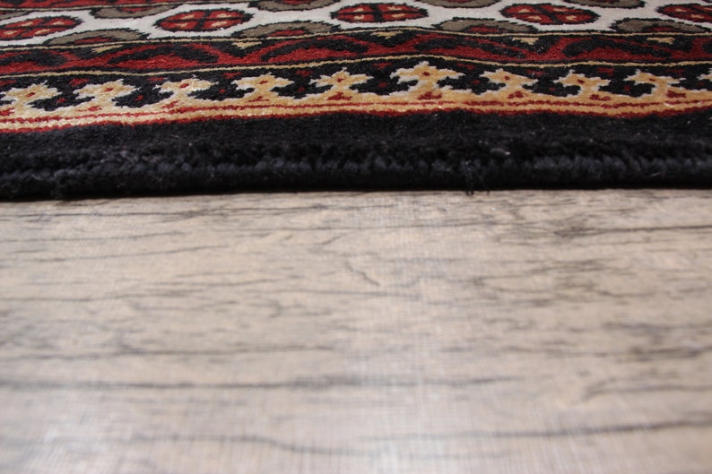 May include: Close-up of the bottom edge of a black rug with a red, white, and gold patterned border. The rug is lying on a light brown wooden floor.