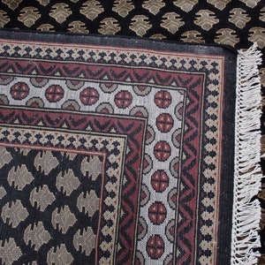 May include: A black and brown patterned rug with a white fringe. The rug has a geometric design with a central square surrounded by a border.