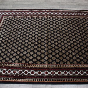 May include: Black area rug with a geometric pattern in beige and white. The rug has a red, white, and beige border.