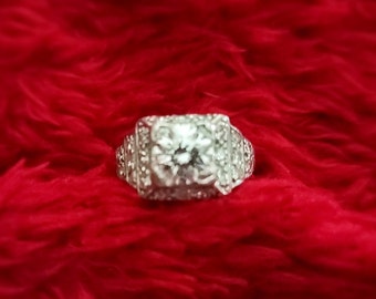 Puede incluir: Un anillo de plata con un gran diamante redondo y claro engastado en un marco cuadrado. El diamante está rodeado de diamantes más pequeños.