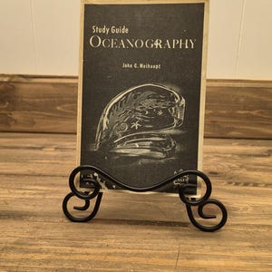 May include: A black metal display stand holding a vintage "Oceanography" study guide. The book has a black and white cover with an illustration of a shell and the title in large, bold letters. The stand has decorative scrollwork.