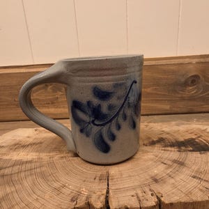 May include: A light gray stoneware mug with a speckled texture and a dark blue hand-painted floral pattern. The mug features a sturdy, curved handle and a cylindrical body with a slightly flared rim, showcasing a classic pottery design.