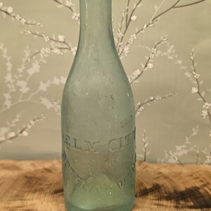 May include: A vintage light blue glass bottle with a rounded top and a narrow neck. The glass features embossed text that reads ELM CITY BOTTLING WORKS in a circular pattern around the body of the bottle, which has a smooth, translucent finish.