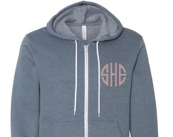 May include: A gray zip-up hoodie with a hood and front pockets. The hoodie features a white zipper and drawstrings, and a circular monogram design on the left chest.