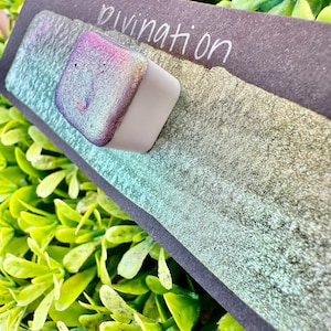 May include: A small square container of iridescent green and purple paint with the word "Divination" written on a black background.
