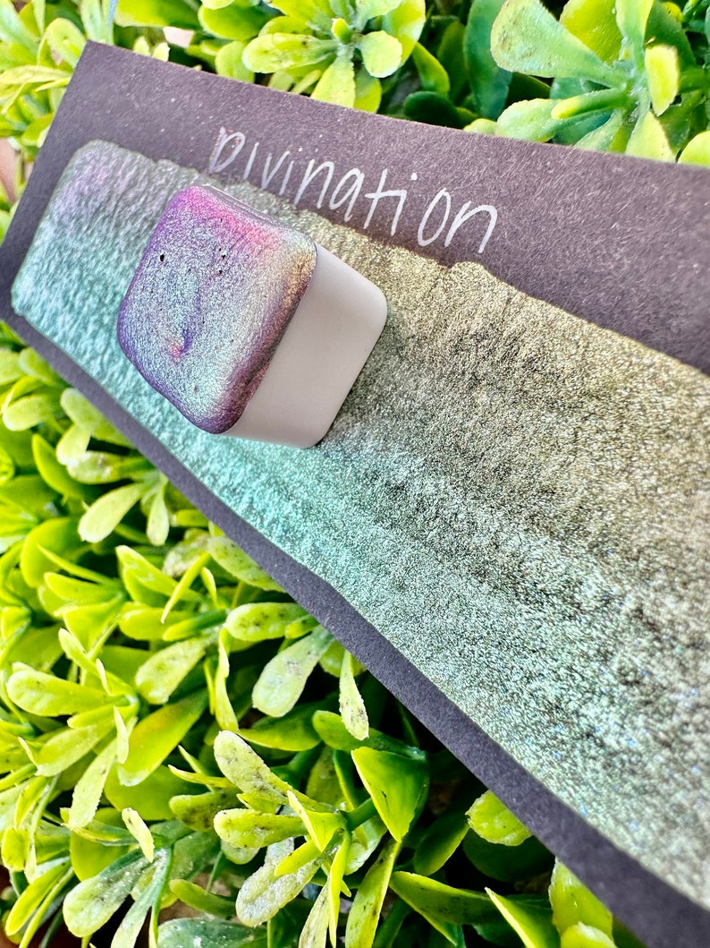 May include: A small square container of iridescent green and purple paint with the word "Divination" written on a black background.