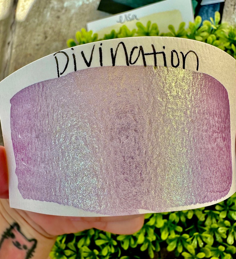 May include: A close-up of a sample of iridescent purple and gold paint with the word "Divination" written above it.