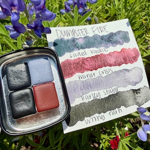 Op de afbeelding: Een klein metalen doosje bevat vier vierkante aquarelverfkoekjes in zwart, grijs en rood. Een kaart ernaast toont aquarelkleurstalen met de tekst "DUMPSTER FIRE" en kleurnamen als "faded hoodie" en "wing turn". De afbeelding is geplaatst tegen een achtergrond van groen gebladerte en paarse bloemen.
