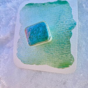 May include: A teal green and iridescent soap bar on a white and green watercolor paper background.