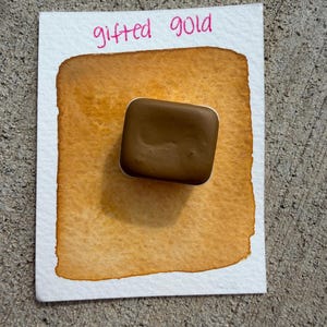 May include: A watercolor swatch on white paper with the words "gifted gold" in pink. The swatch is a gradient of gold and brown tones, with a solid brown square of paint in the center.